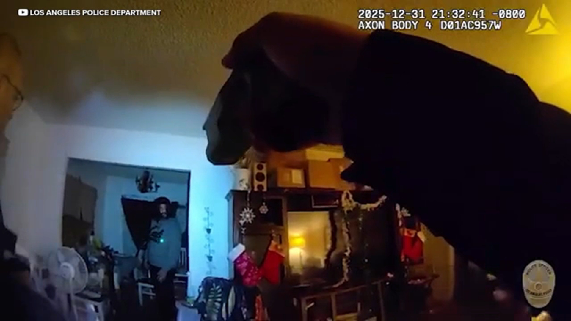 A Los Angeles Police Department officer standing on a city sidewalk at night wearing a body camera and uniform, with streetlights in the background, news photography style
