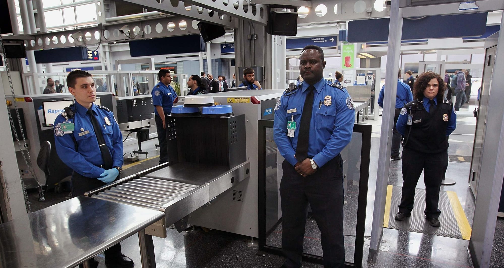 A TSA officer standing at an airport security checkpoint lane with travelers and screening equipment in the background, candid news photography style