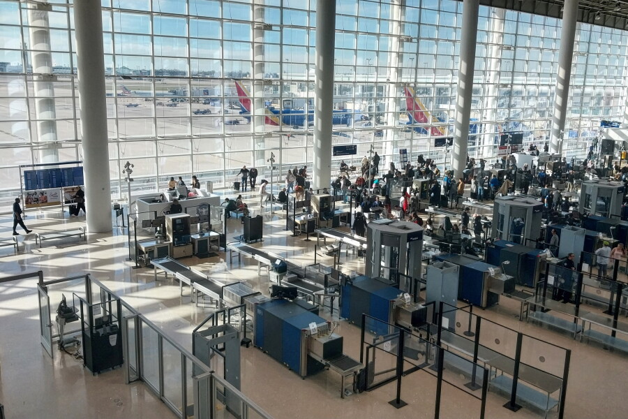 A TSA travel document checker station at an airport checkpoint entrance, with passengers queued and an officer positioned at the ID-check podium