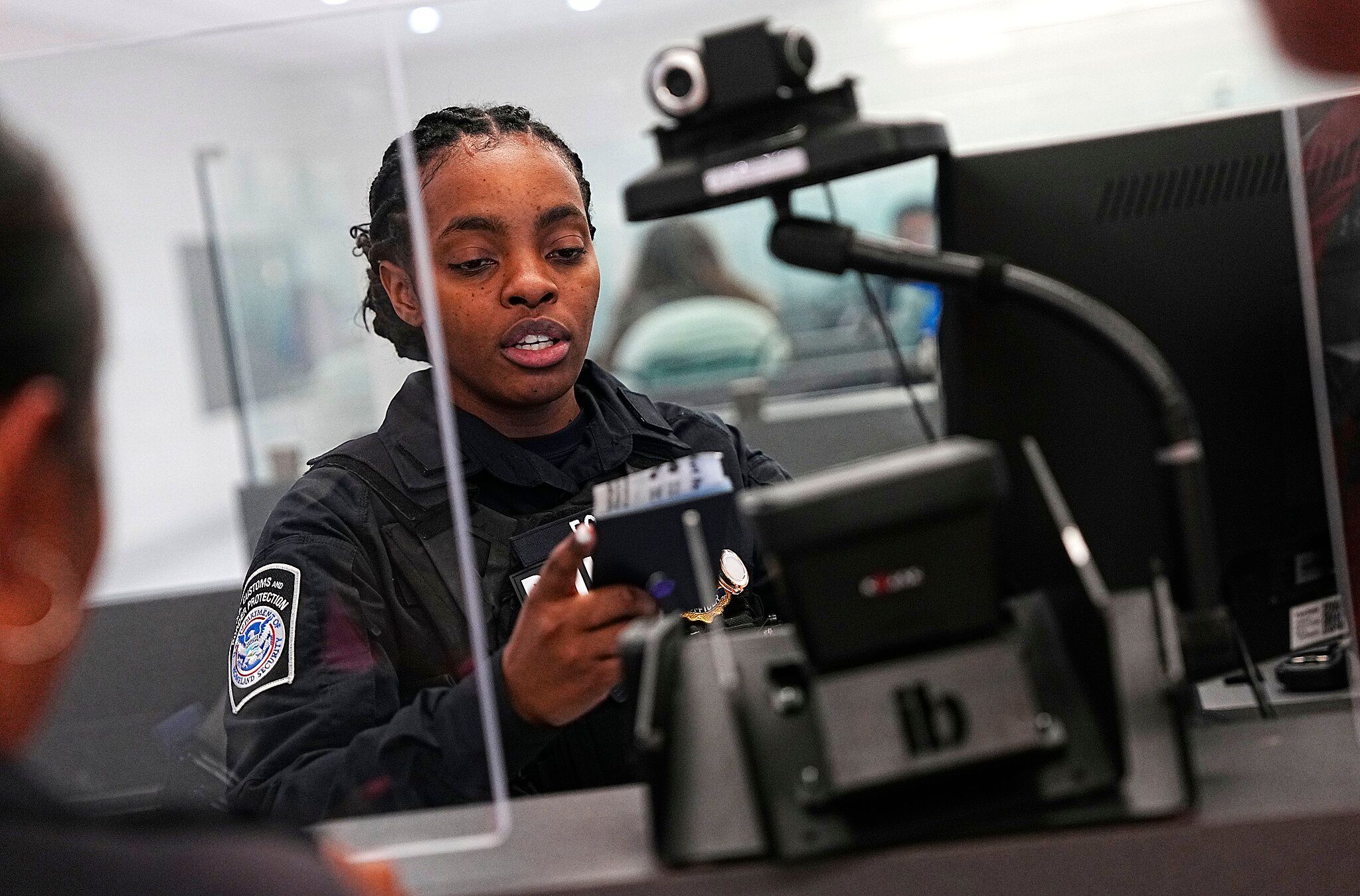 A U.S. Customs and Border Protection officer holding a smartphone while speaking with a traveler in an inspection area