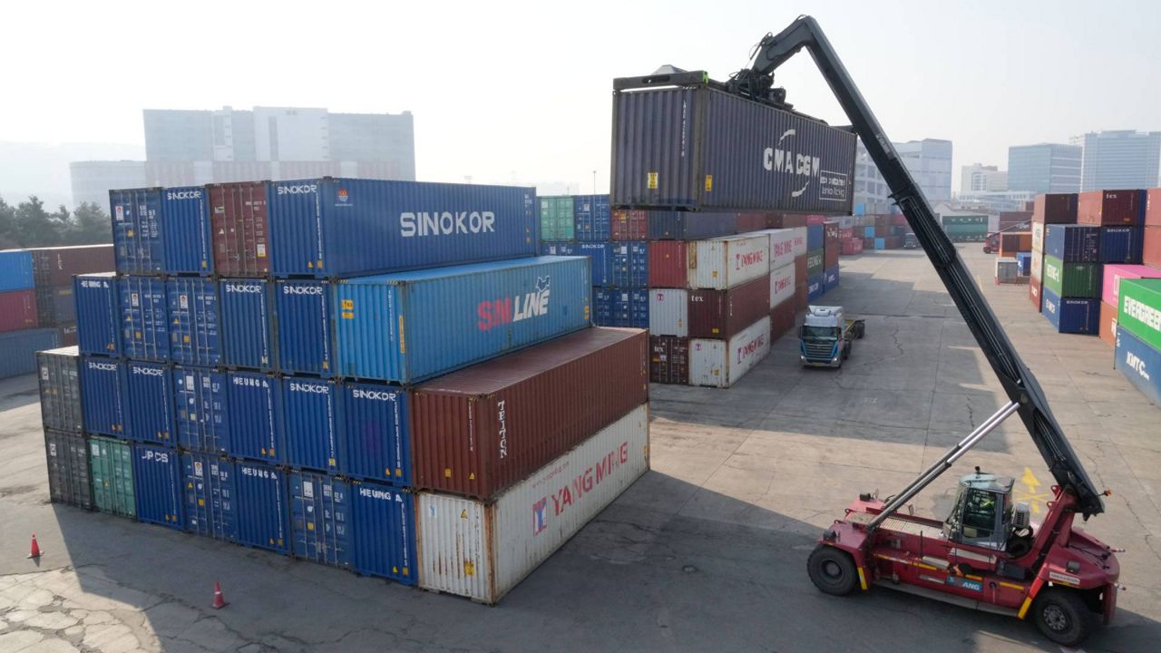 A U.S. Customs officer standing near pallets of imported goods at a cargo terminal while workers move freight in the background, news photography style