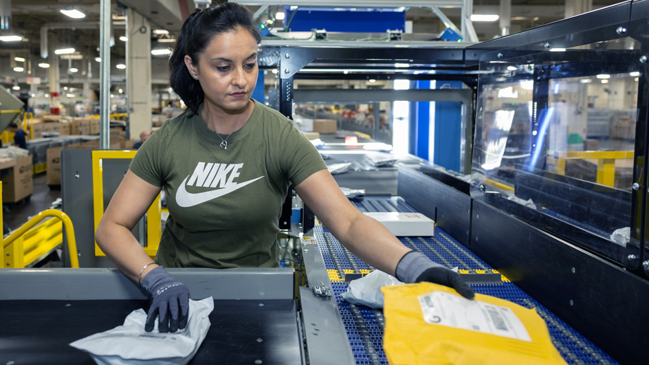 A U.S. Postal Service employee scanning outgoing parcels on a conveyor belt inside a large mail processing facility, news photography style
