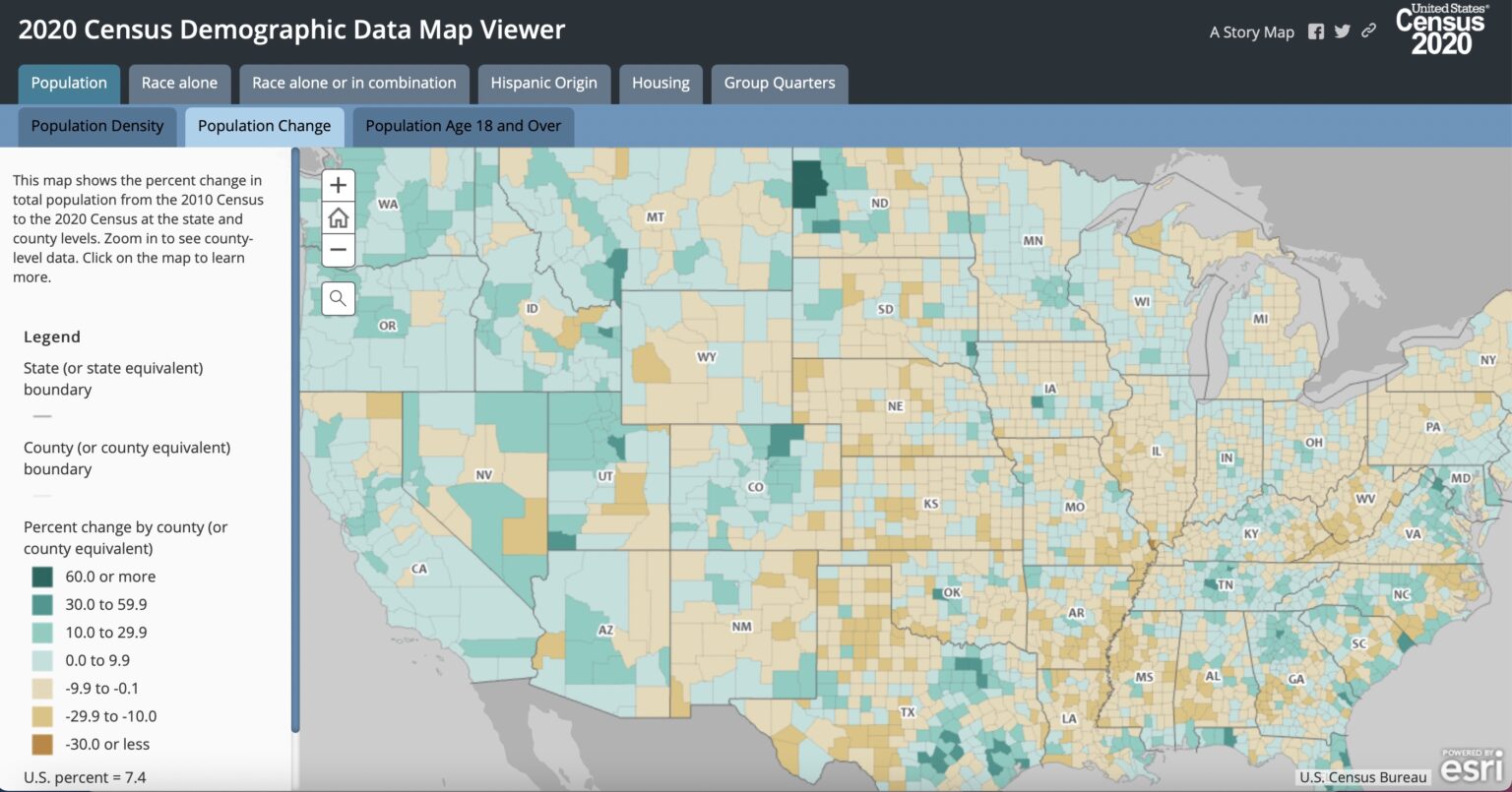 A U.S. census enumerator wearing a government ID badge standing on a residential sidewalk and speaking with a homeowner at the front door, documentary news photo style