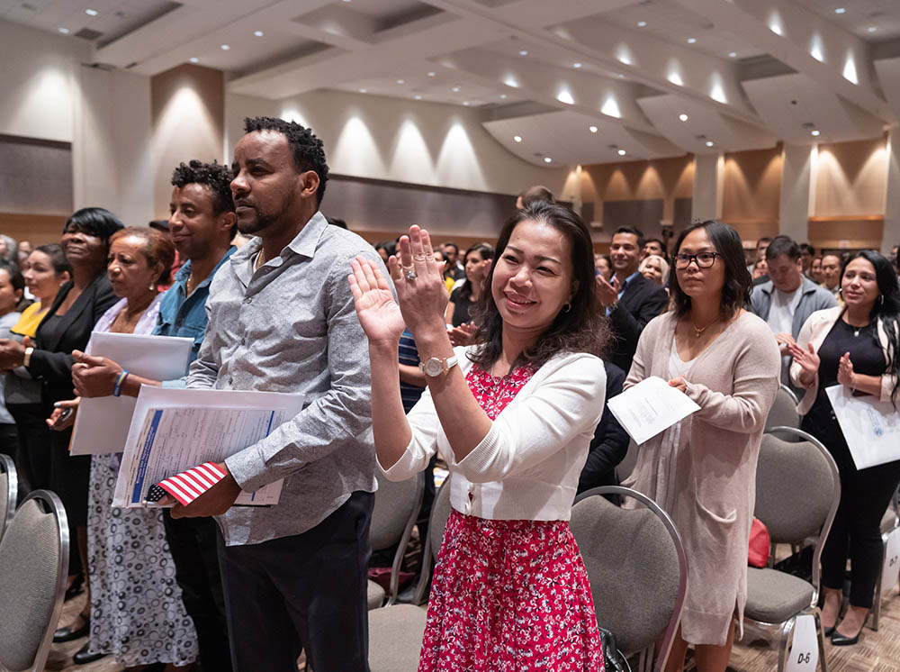 A USCIS naturalization ceremony in a community center, with families watching as new citizens hold small American flags during the oath moment, documentary photograph