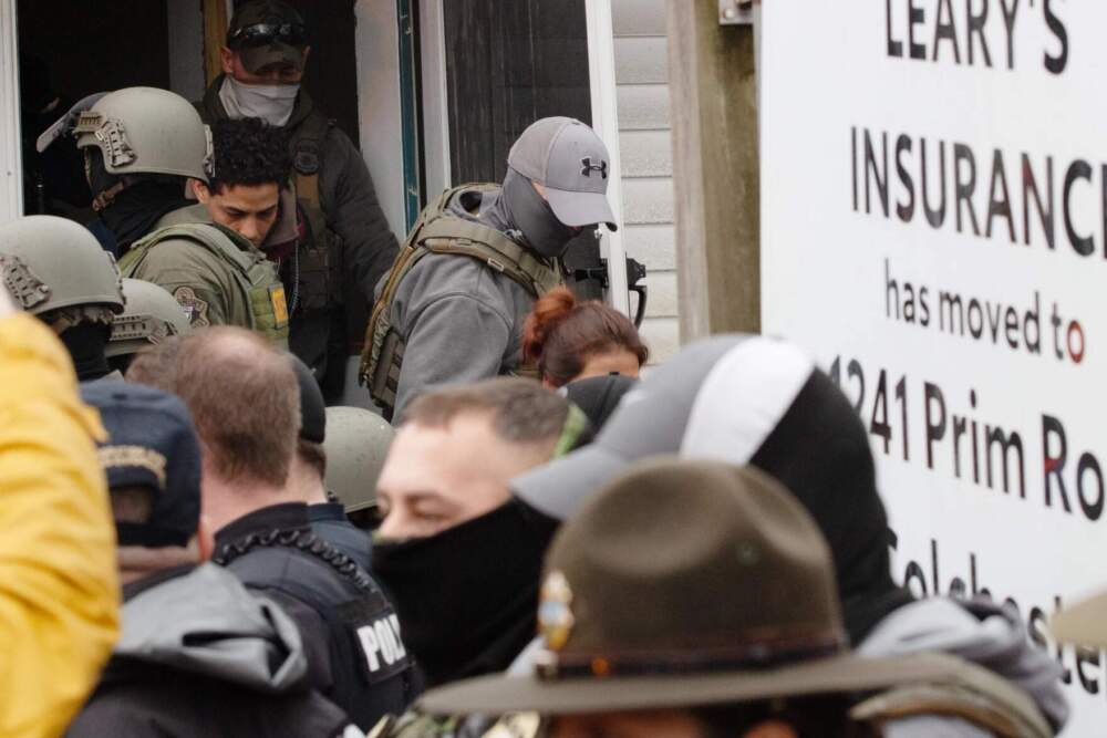A Vermont state capitol hallway during a protest, with a state police officer lifting a seated demonstrator to her feet while other protesters sit linked arm in arm nearby, realistic news photography