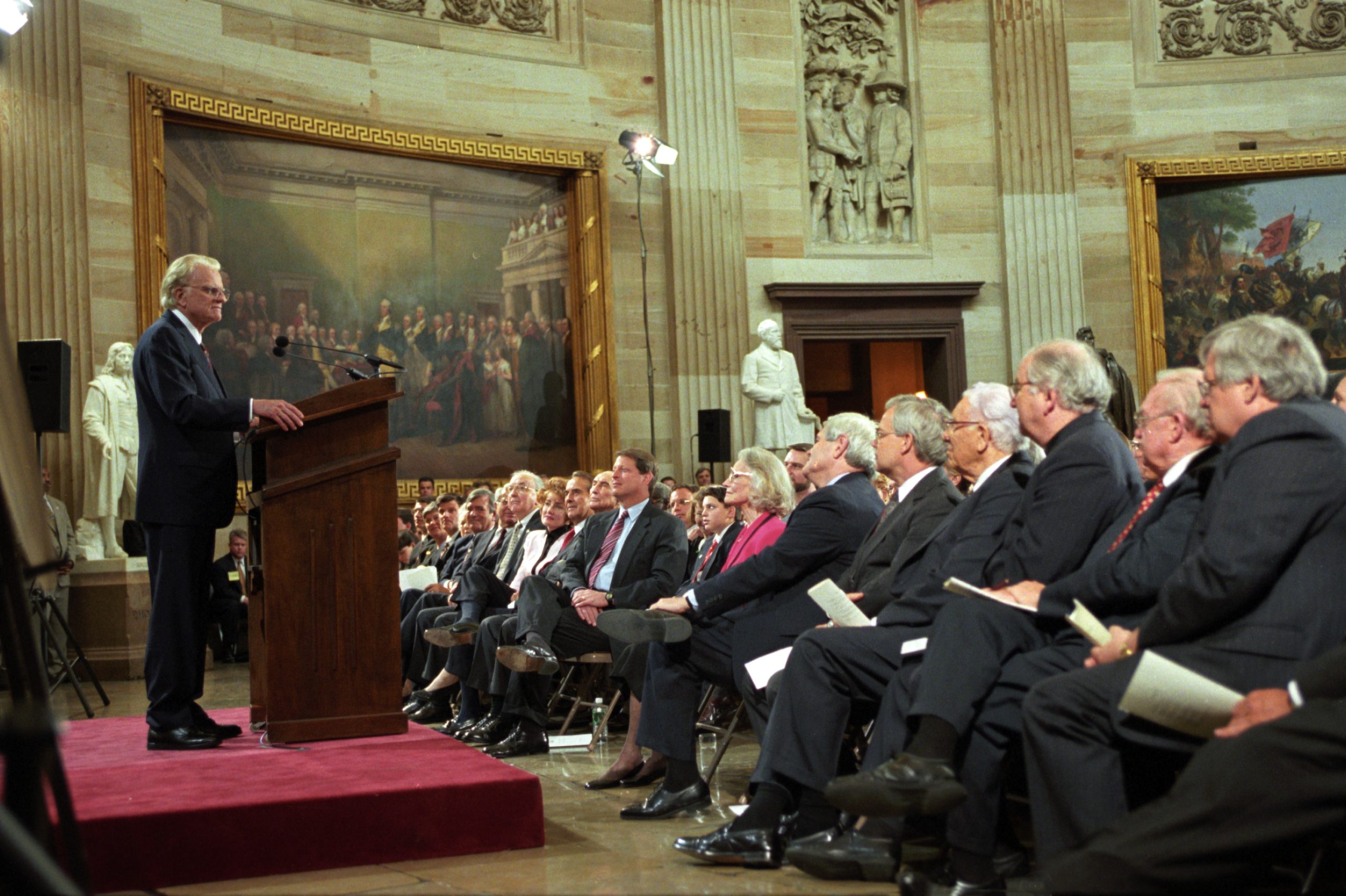 A bill signing ceremony in a formal government room with a wooden desk, a newly signed document on the desk, and American flags in the background, news photography style