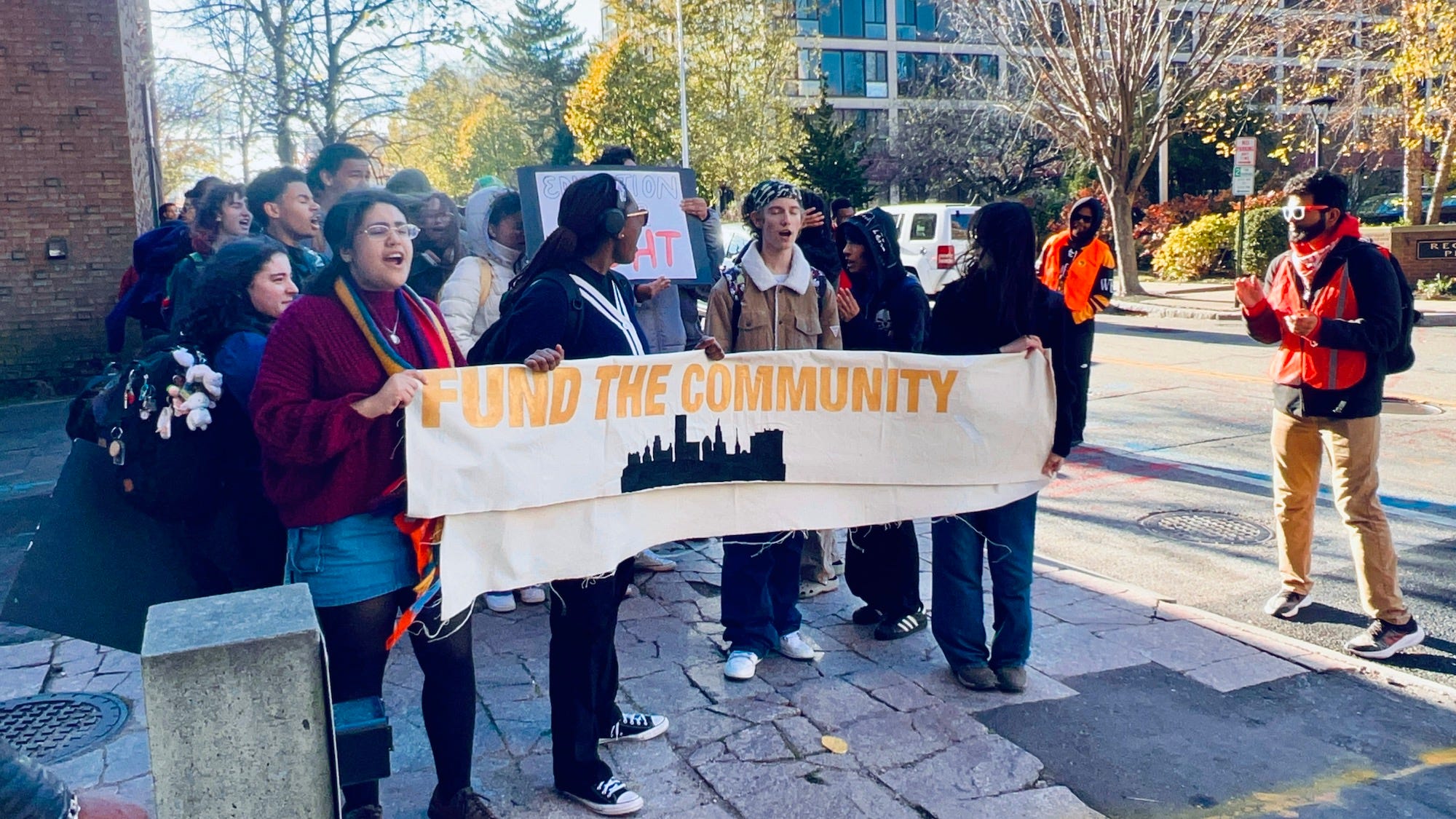 A candid photograph of a public high school hallway with students walking between classes, some wearing backpacks and school spirit clothing, natural indoor lighting