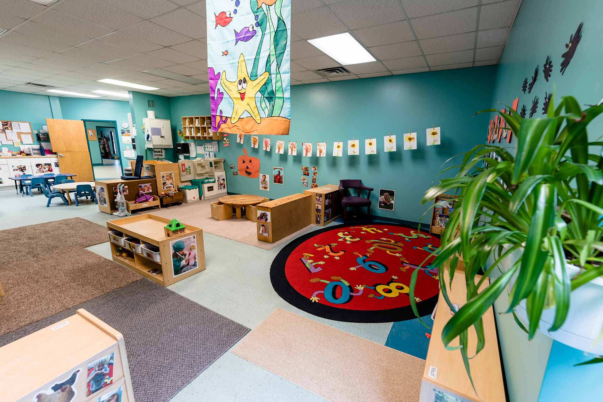 A child care classroom with small tables, colorful toys, and a caregiver organizing materials while children play in the background, realistic documentary photo