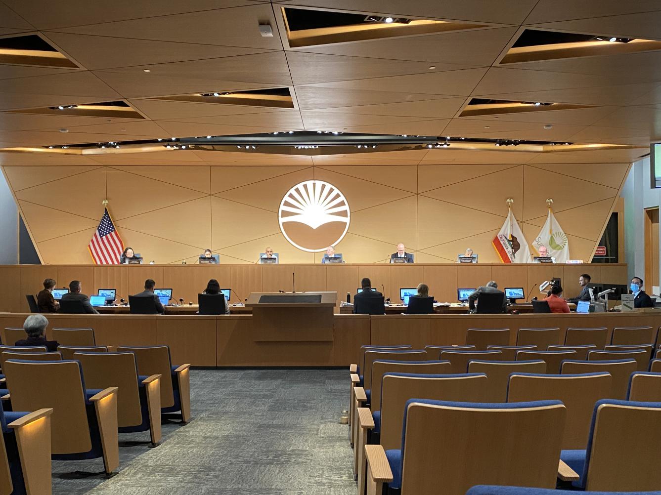 A city council chamber during a public meeting with council members seated at a dais and attendees in the gallery, documentary news photo style
