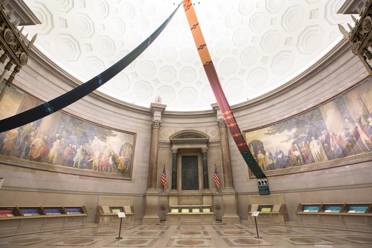 A close, documentary-style photograph of the Fourteenth Amendment page displayed inside the National Archives Rotunda in Washington, DC, with visitors blurred in the background