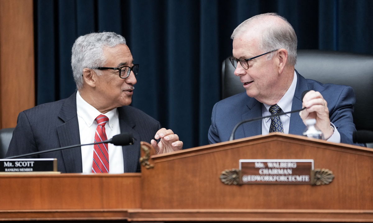 A close-up photograph of a House clerk at a desk in the chamber handling stacks of paper during legislative proceedings, documentary news photography style