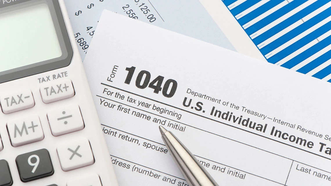 A close-up photograph of a calculator on top of federal tax forms with a pen and reading glasses on a wooden desk, shallow depth of field