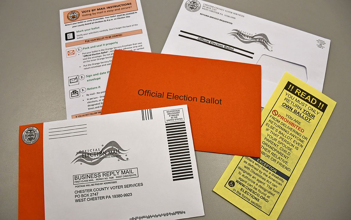 A close-up photograph of a voter’s hands placing a sealed mail ballot envelope on a kitchen table next to a pen and folded election instructions, natural indoor light, documentary news style