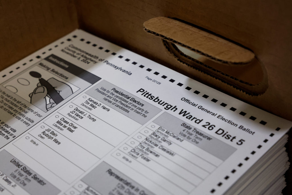 A close-up real photograph of a hand placing a completed presidential ballot into a secure ballot box at a polling place in Pennsylvania, documentary news photography style
