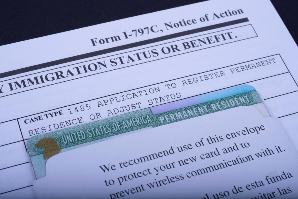 A close view of a person’s hands holding a stack of immigration paperwork on a desk, with a pen and an open folder beside it, documentary news photography style