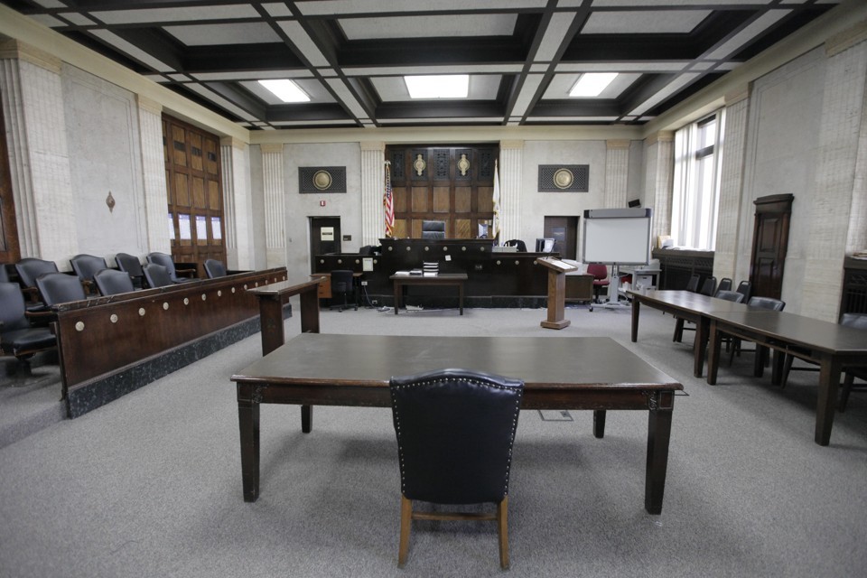 A criminal courtroom scene with a defense attorney seated beside a defendant at the defense table while a prosecutor stands near the jury box, realistic news photography style