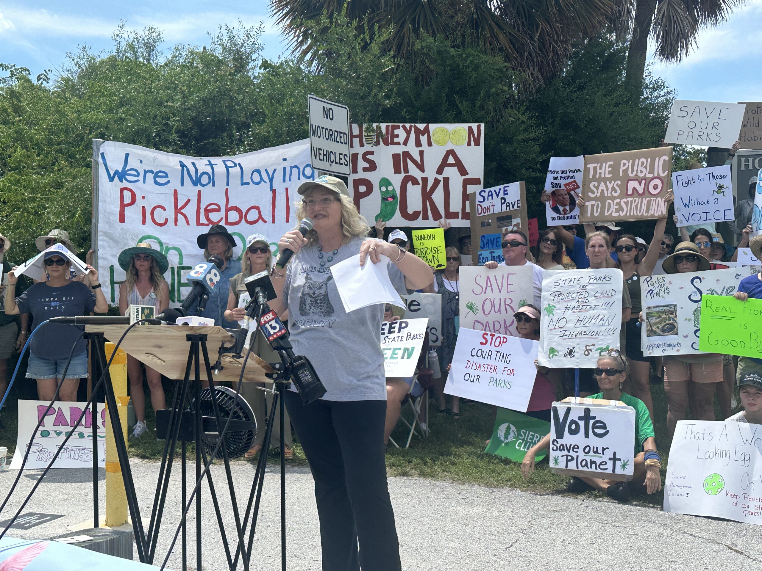 A crowd gathered in a public park for a rally, holding handmade signs and listening to a speaker on a small platform, real photograph style