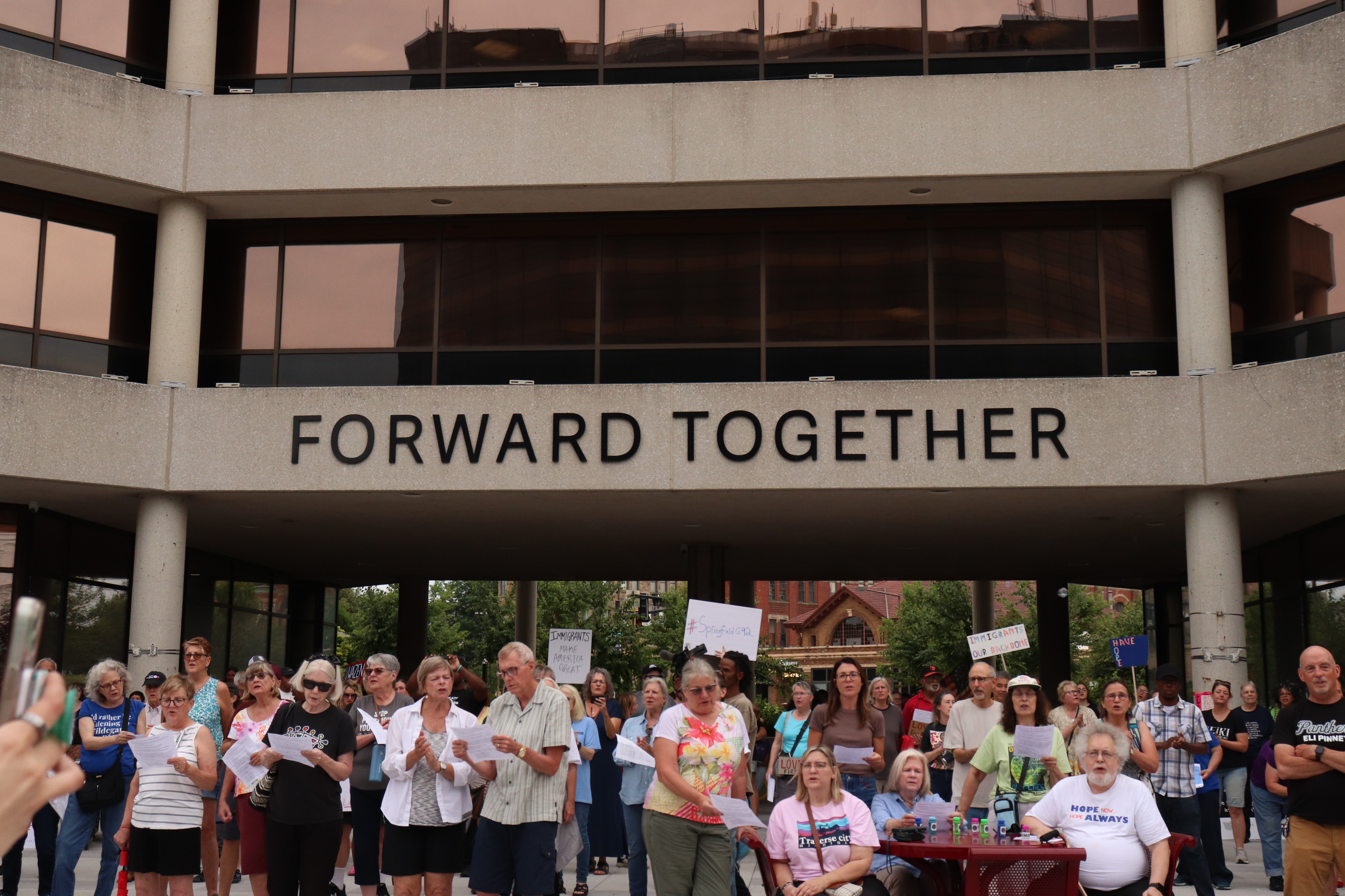 A crowd gathered in the stone courtyard outside Springfield City Hall during an evening vigil, with people holding signs and listening to speakers