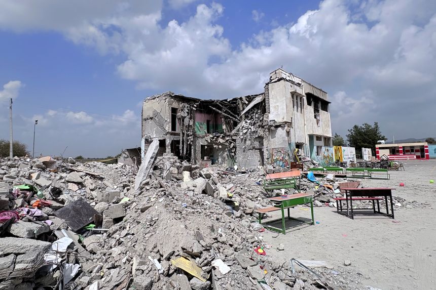 A damaged school building with broken walls and debris scattered in front, photographed in daylight by a news photographer