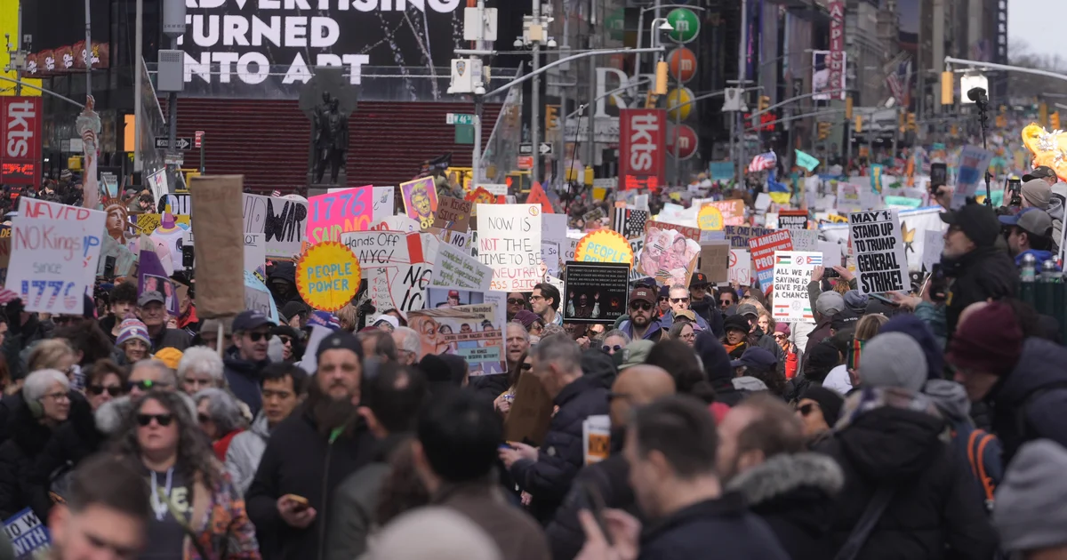 A dense crowd marching through a New York City street during a daytime No Kings protest, people holding handmade signs, news photography style