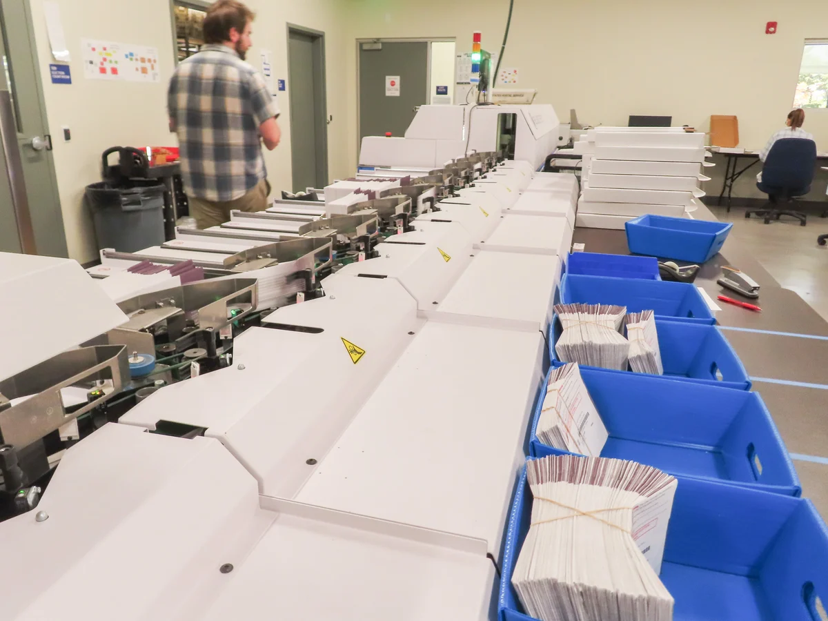 A documentary-style photograph inside a county election office where election workers sort outgoing ballot packets on long tables under bright overhead lighting