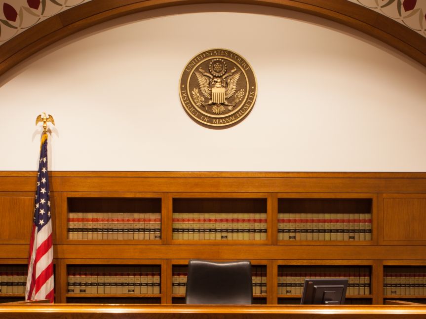 A federal district court courtroom during a hearing, with counsel tables in the foreground and the judge’s bench in the background, photographed from the public gallery