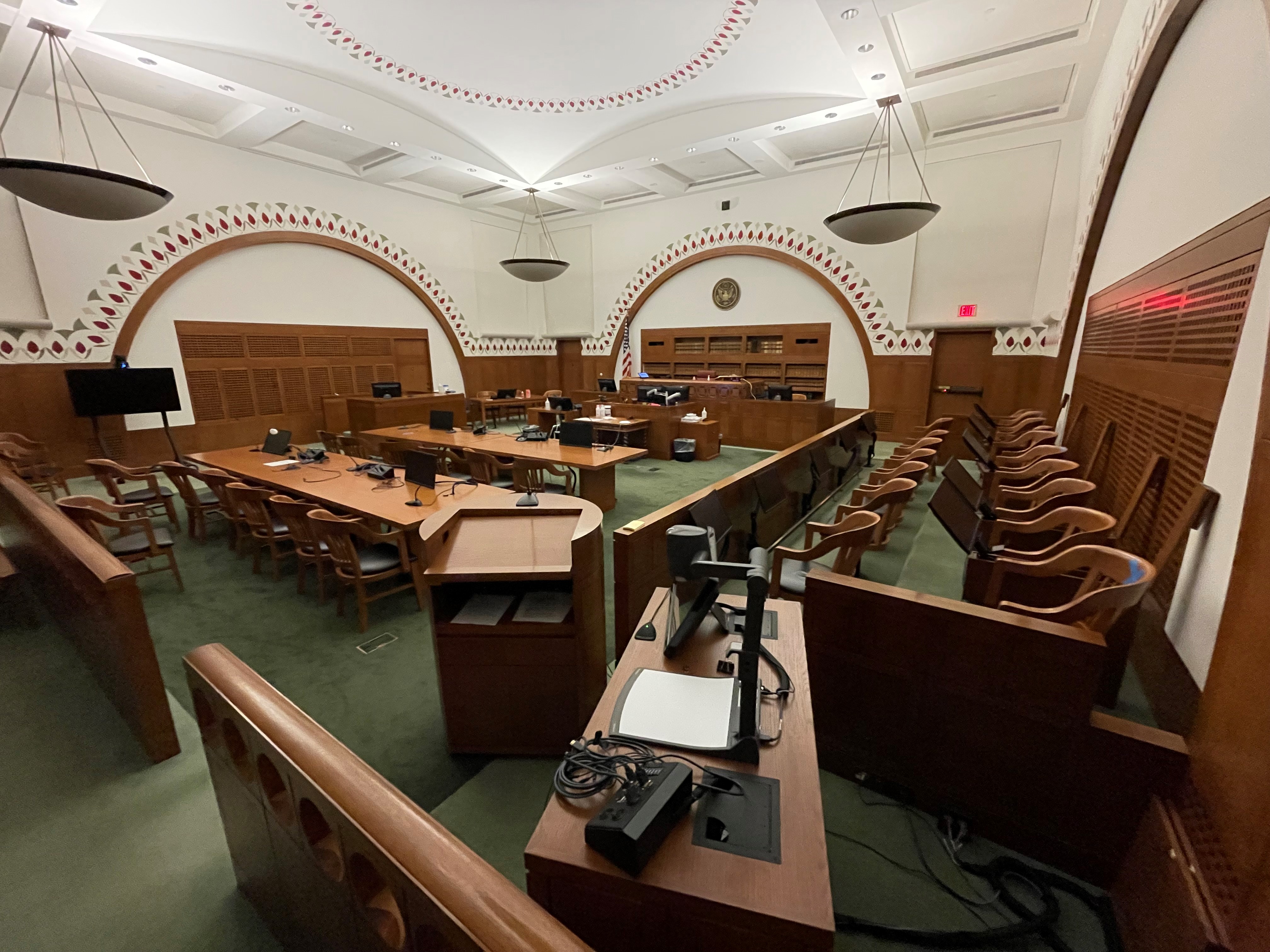A federal district courtroom interior with the judge’s bench, counsel tables, and the U.S. flag behind the bench, empty before proceedings, news photography style
