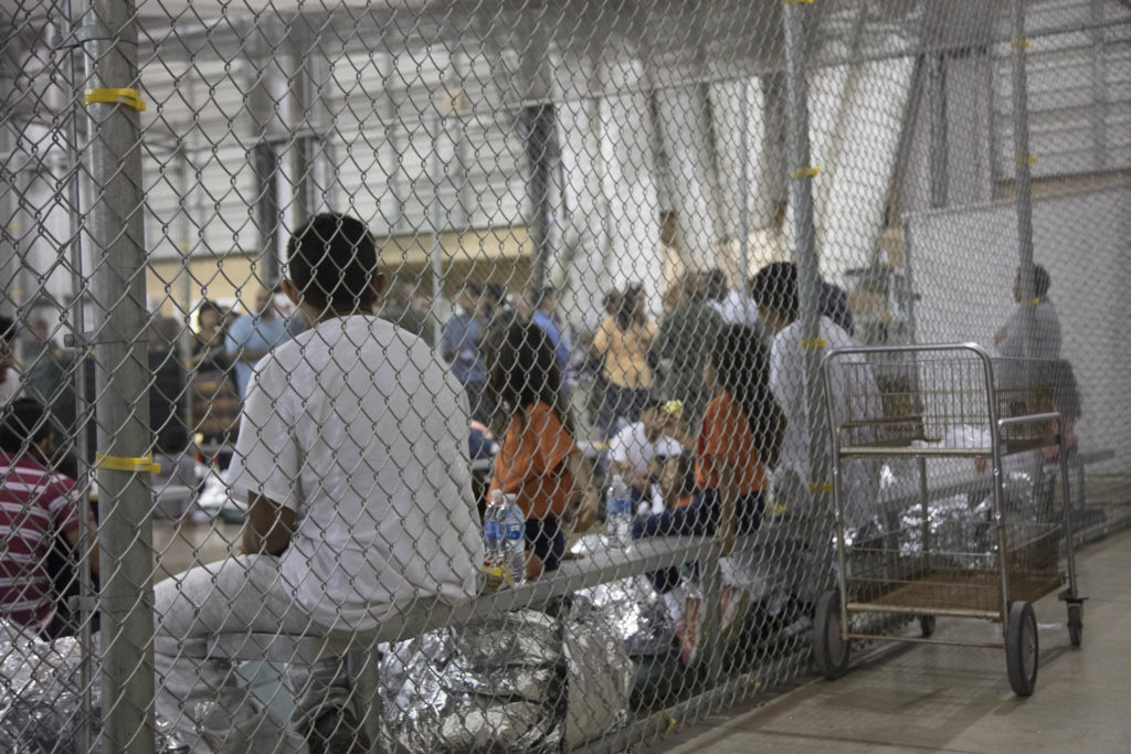 A federal immigration detention facility exterior with a chain link fence and a U.S. flag flying nearby, shot in daylight, news photography style