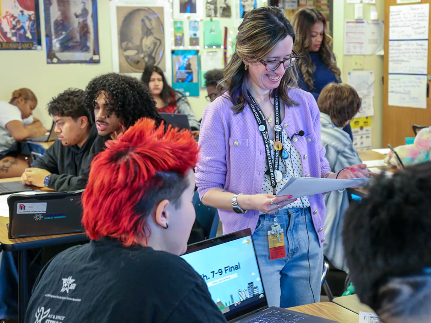A high school civics classroom where students read primary-source constitutional debates on laptops while a teacher points to a projected document, news photography style