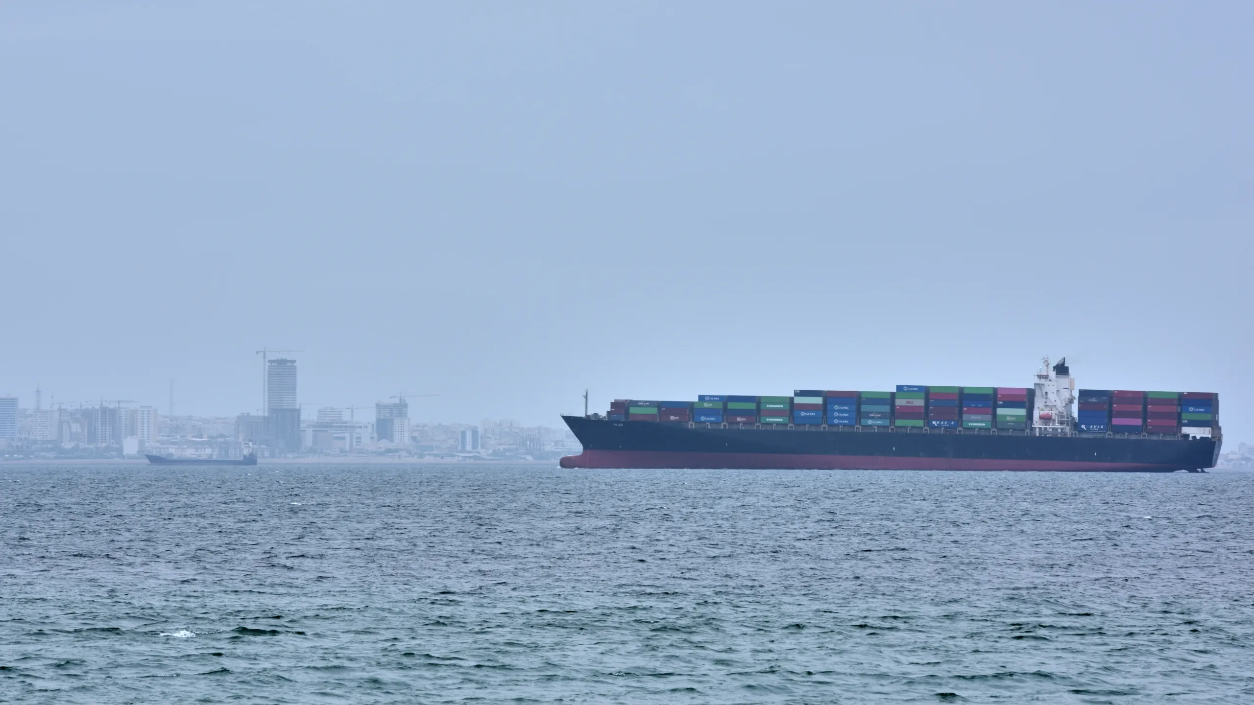 A large commercial cargo ship transiting a narrow sea lane with distant naval vessels on the horizon under clear skies, news photography style