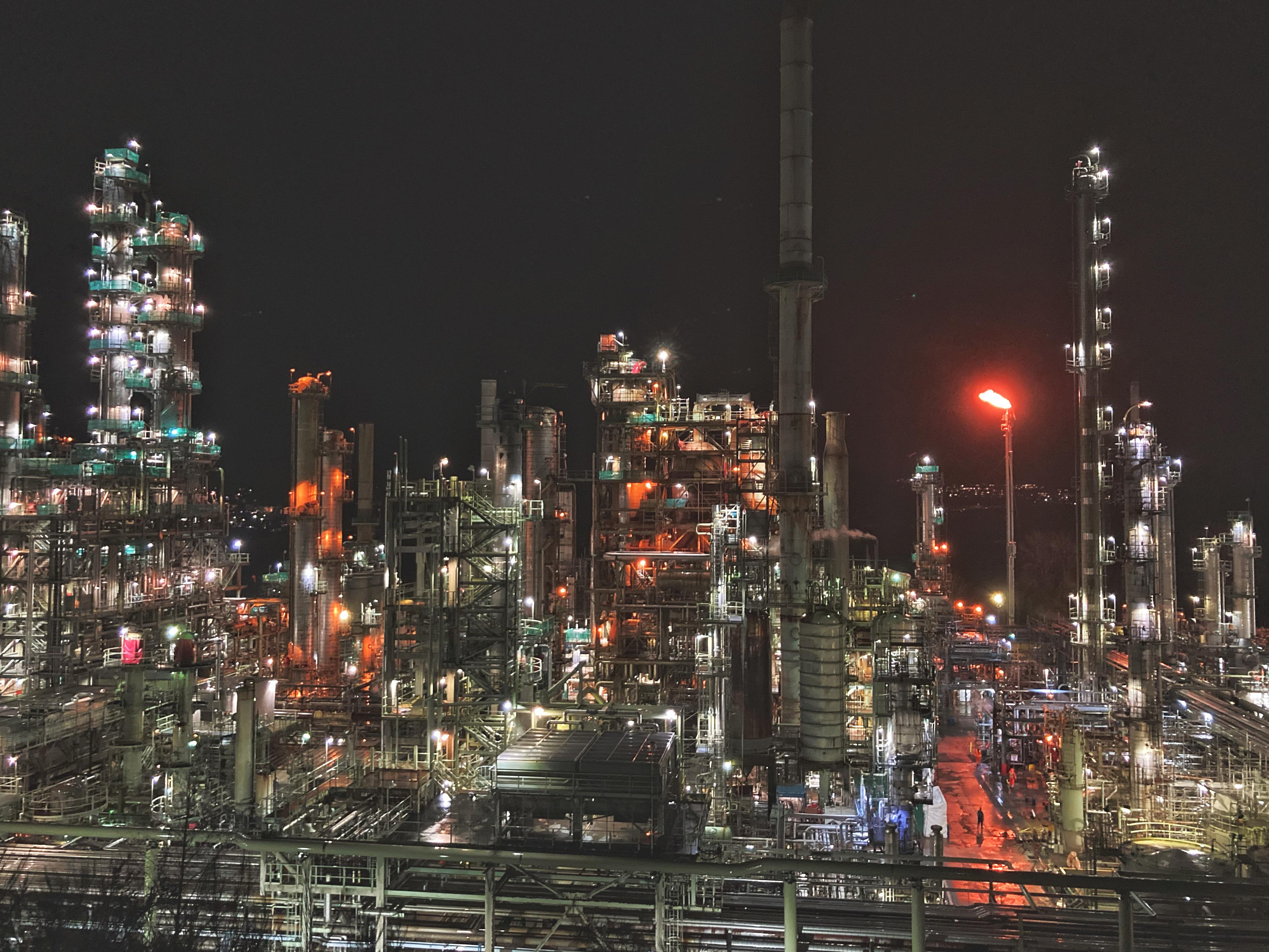 A large oil refinery at night with bright industrial lights, pipelines, and storage tanks visible under a dark sky, realistic news photography