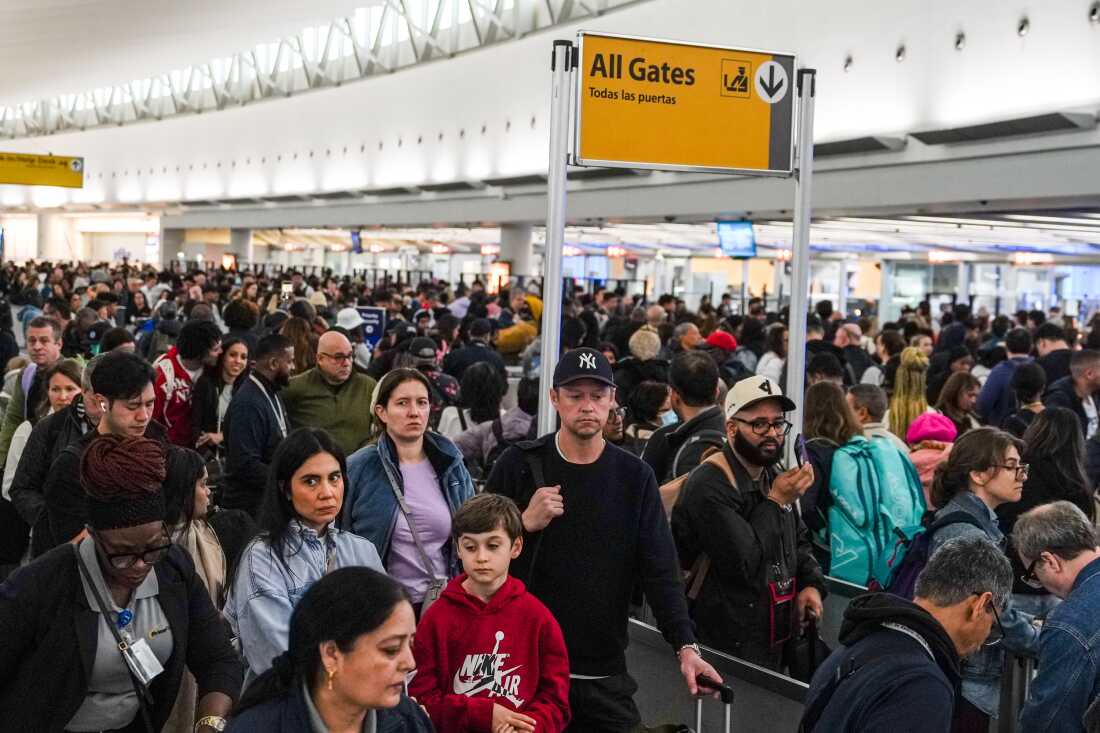 A long TSA security line inside John F. Kennedy International Airport with travelers waiting in stanchioned lanes and screening equipment visible ahead, news photography style