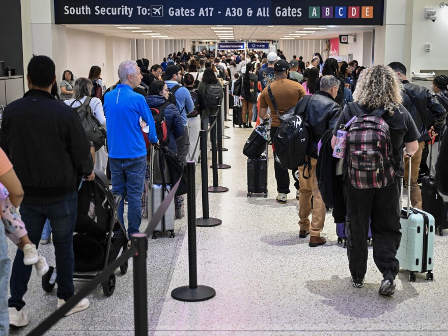 A long TSA security line stretching through an airport terminal with passengers waiting and a TSA checkpoint visible in the background, news photography style