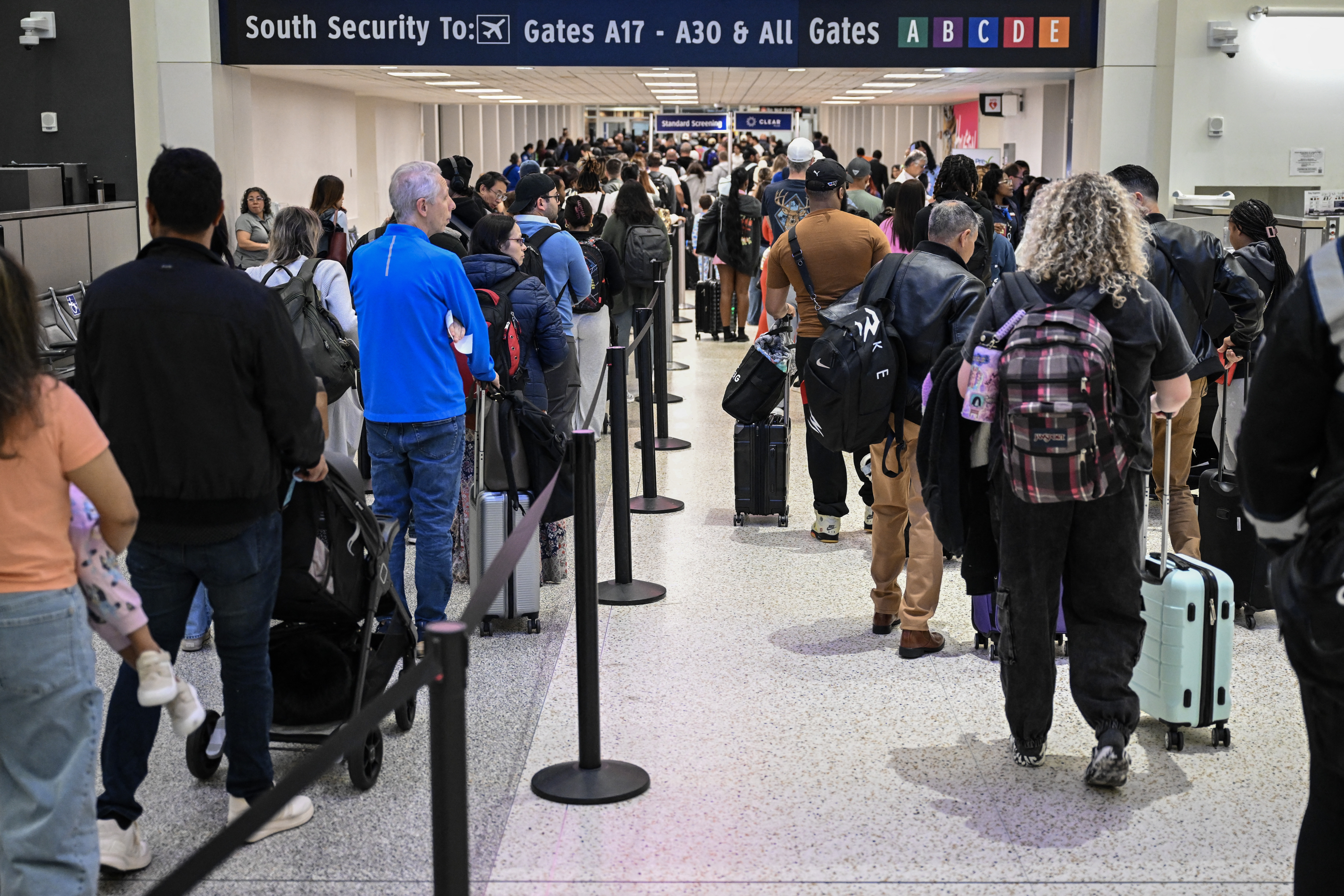 A long line of travelers waiting at a TSA security checkpoint inside a busy American airport terminal, with stanchions and overhead signage visible, photorealistic news photography