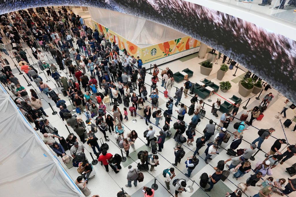 A long line of travelers waiting at a TSA security checkpoint inside a busy U.S. airport terminal, with stanchions and overhead departure boards in the background, news photography style