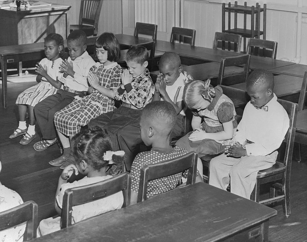 A mid-1960s public school classroom in the Southern United States with students of different races seated together while a teacher stands at the front, documentary news photography style