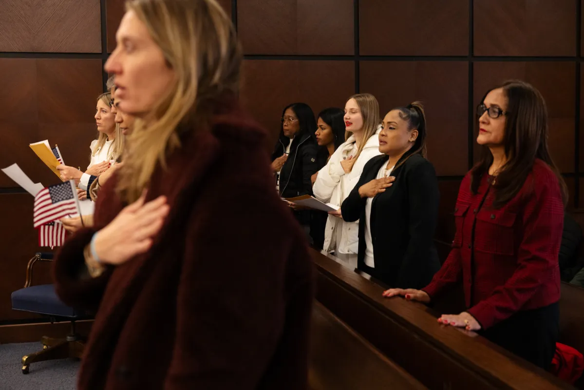 A naturalization ceremony with new citizens standing and holding small American flags inside a federal courthouse ceremonial room, news photography style