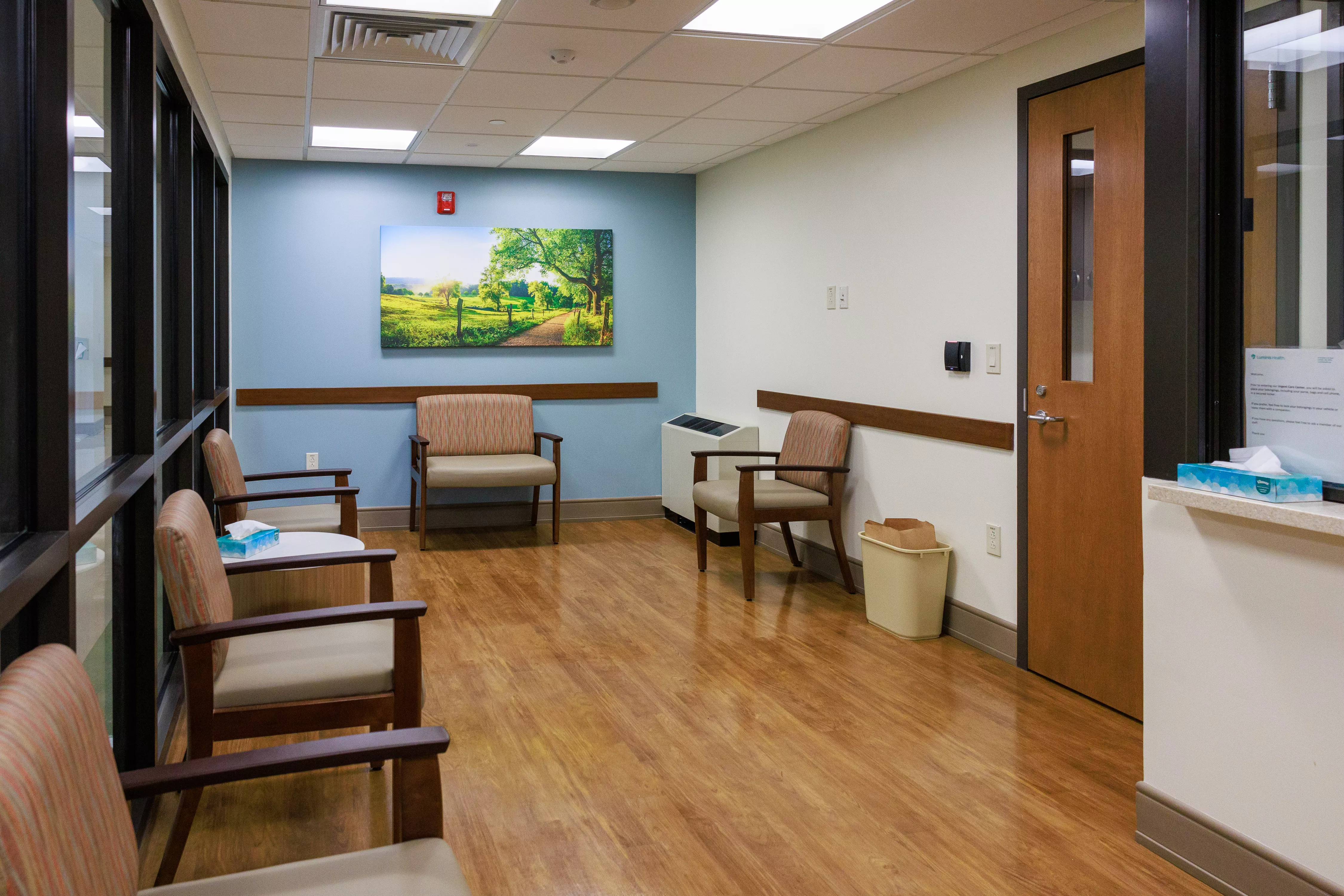 A news-style photograph of a community health clinic waiting room with patients seated quietly and an intake window in the background