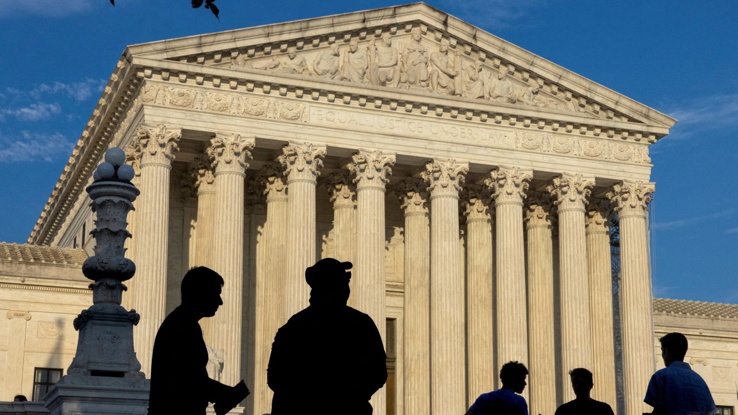 A news-style photograph of the United States Supreme Court building in Washington, DC on a bright summer day, with visitors gathered on the front steps