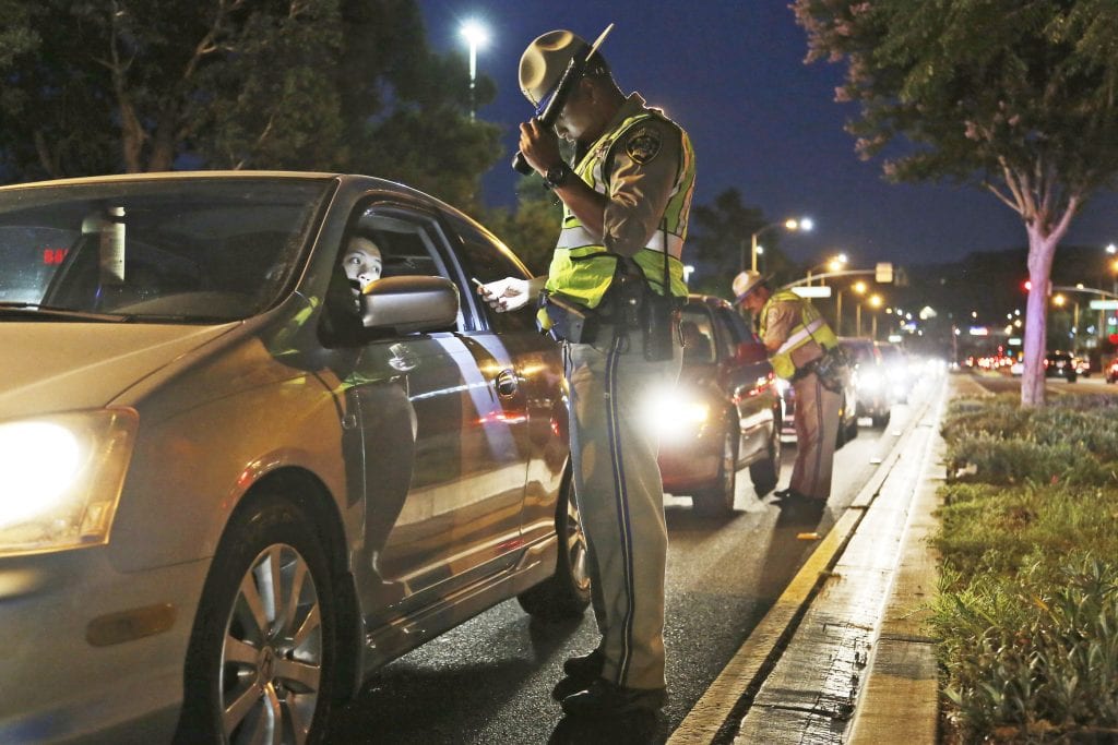 A nighttime sobriety checkpoint with uniformed officers speaking to drivers at a line of traffic cones under bright patrol lights on a city street, news photography style