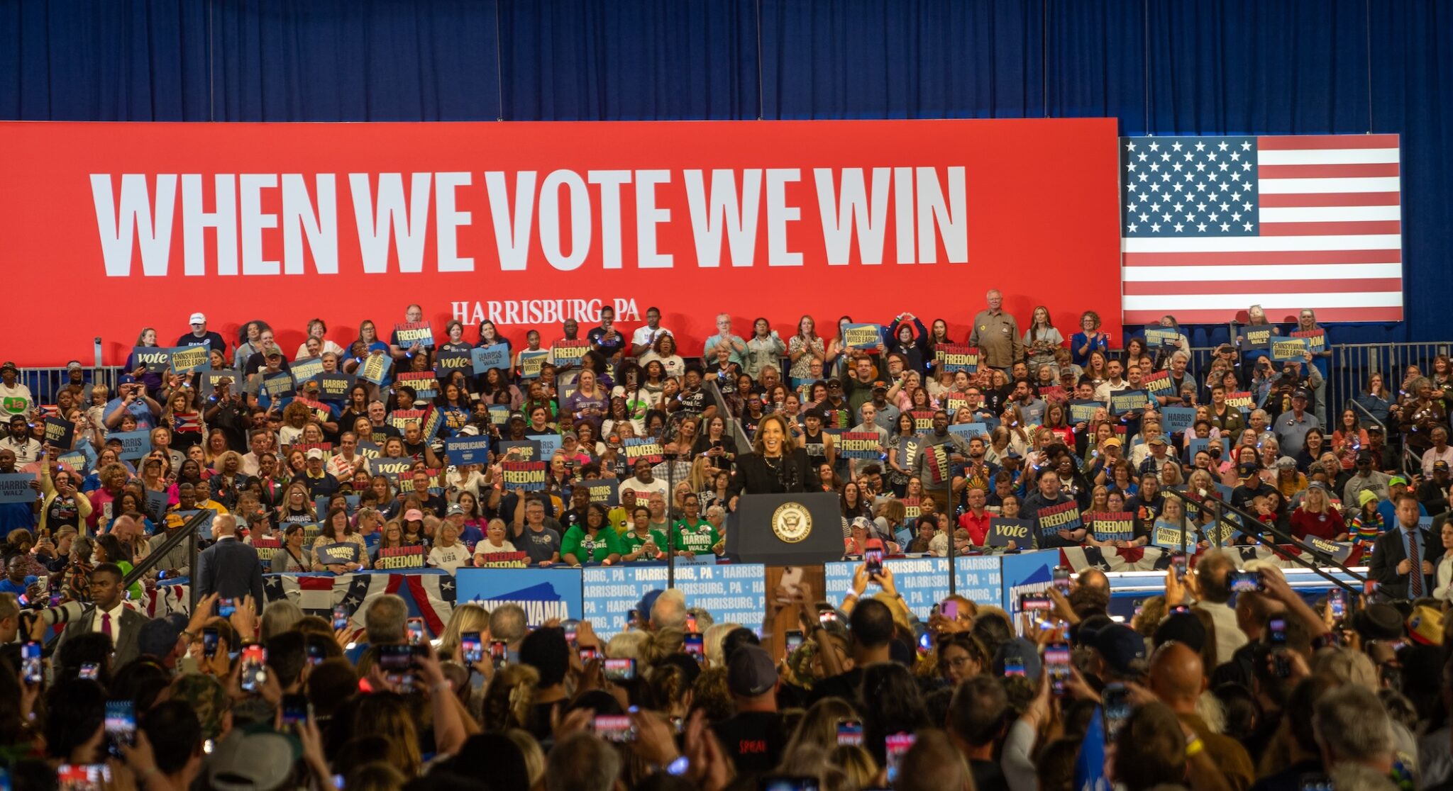A photograph of a crowded outdoor campaign rally with attendees holding American flags and looking toward a stage in the distance