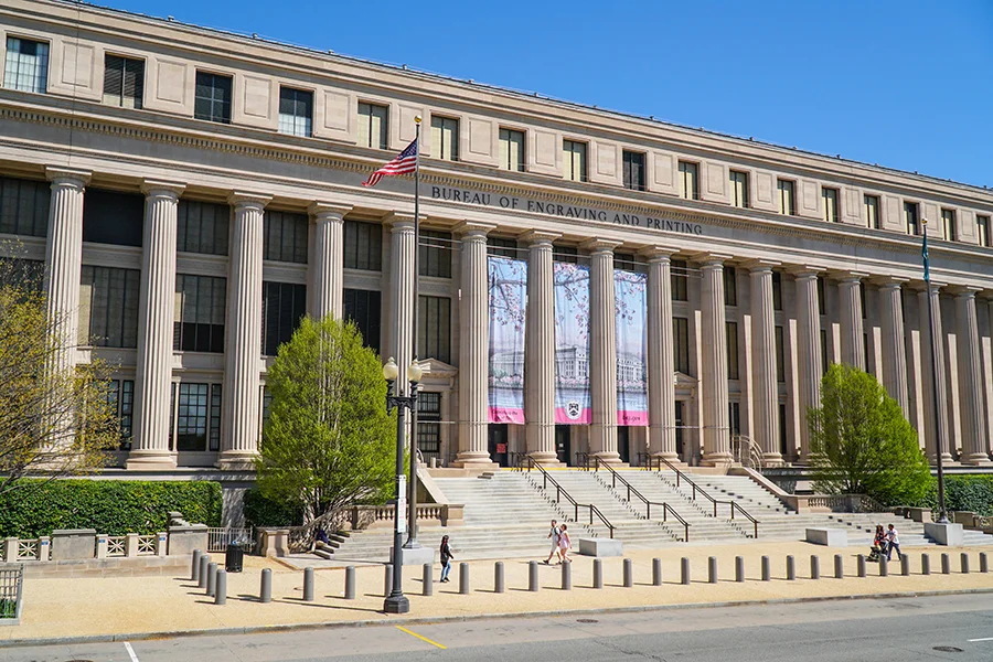 A photograph of the Bureau of Engraving and Printing building in Washington, D.C. on a clear day, with the facade centered and a few pedestrians in the foreground, realistic news photography