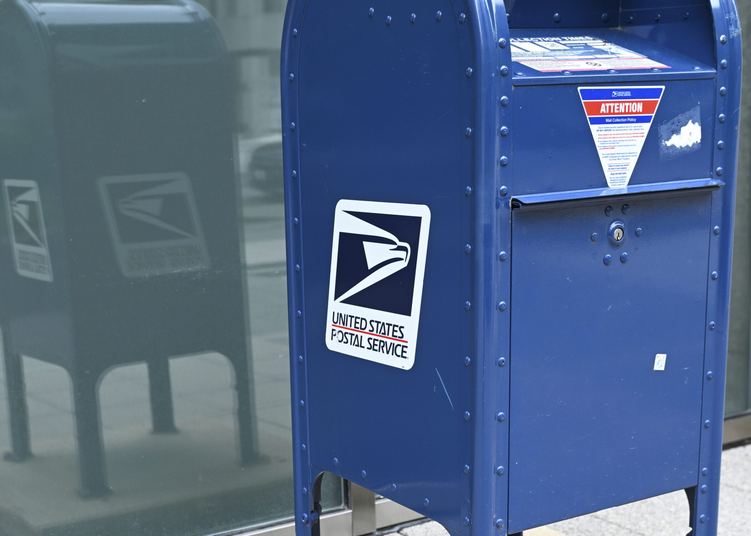 A postal clerk standing behind a counter at a United States post office with shipping supplies in the foreground, documentary news photo style