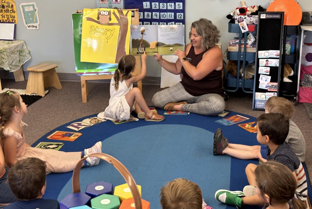 A preschool classroom in Denver with small tables, children’s backpacks on hooks, and a teacher preparing materials before students arrive, realistic news photography style