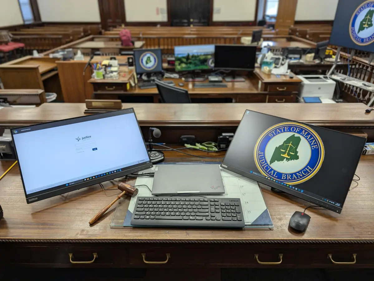 A public-access computer terminal inside a Maine courthouse hallway showing a court docket search screen, with people walking past in the background, news photography style