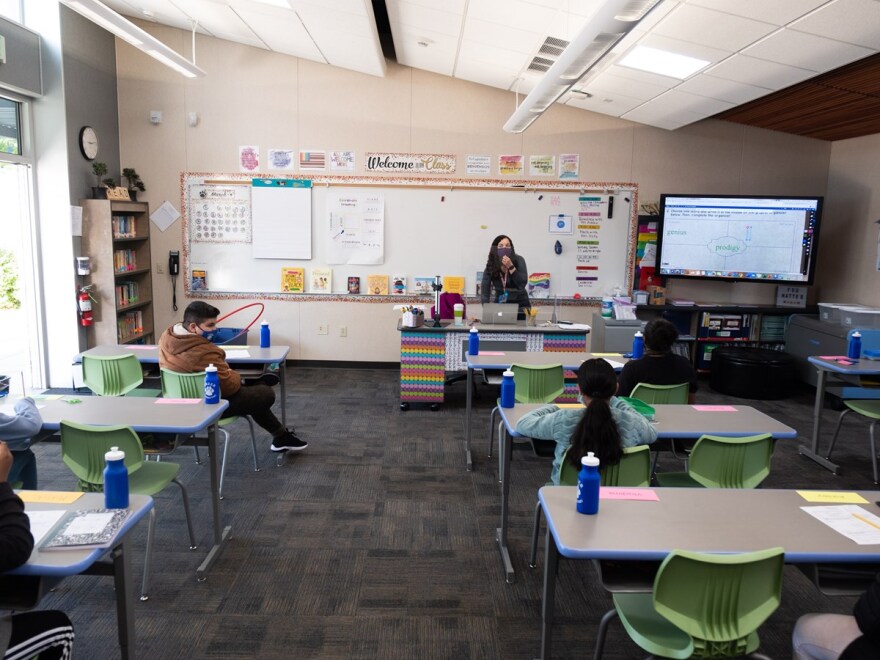 A quiet public school classroom with empty desks facing a whiteboard, a U.S. flag in the corner, and a plain bulletin board on the wall, realistic news photography