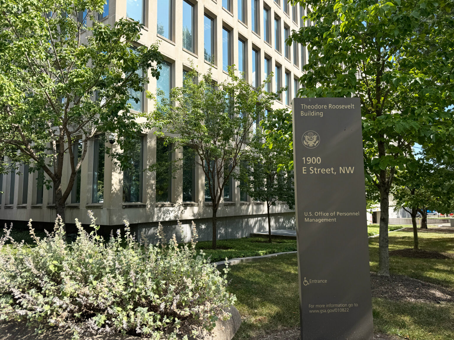 A real photo of a federal agency building entrance with people entering through glass doors and a visible security checkpoint inside, news photography style
