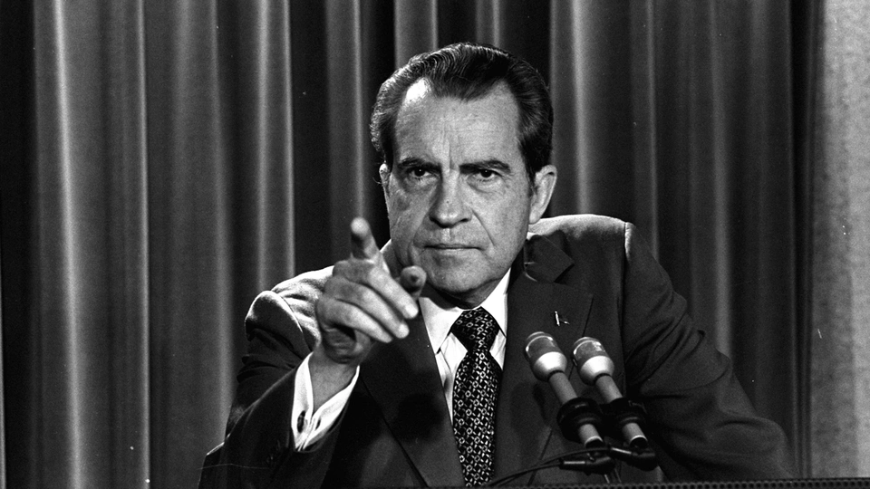 A real photograph of President Richard Nixon speaking at a lectern during a White House press conference in the early 1970s, news photography style