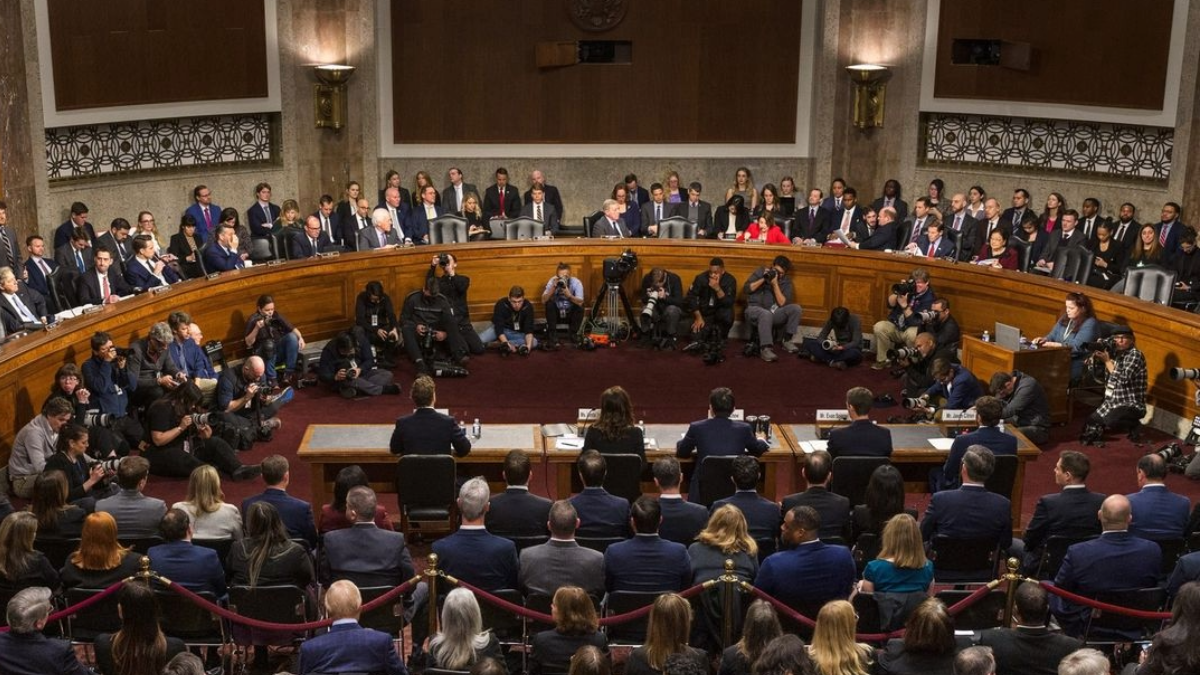 A real photograph of a U.S. Senate committee hearing where a witness sits at a table while senators look on, with microphones and nameplates visible, news photography style