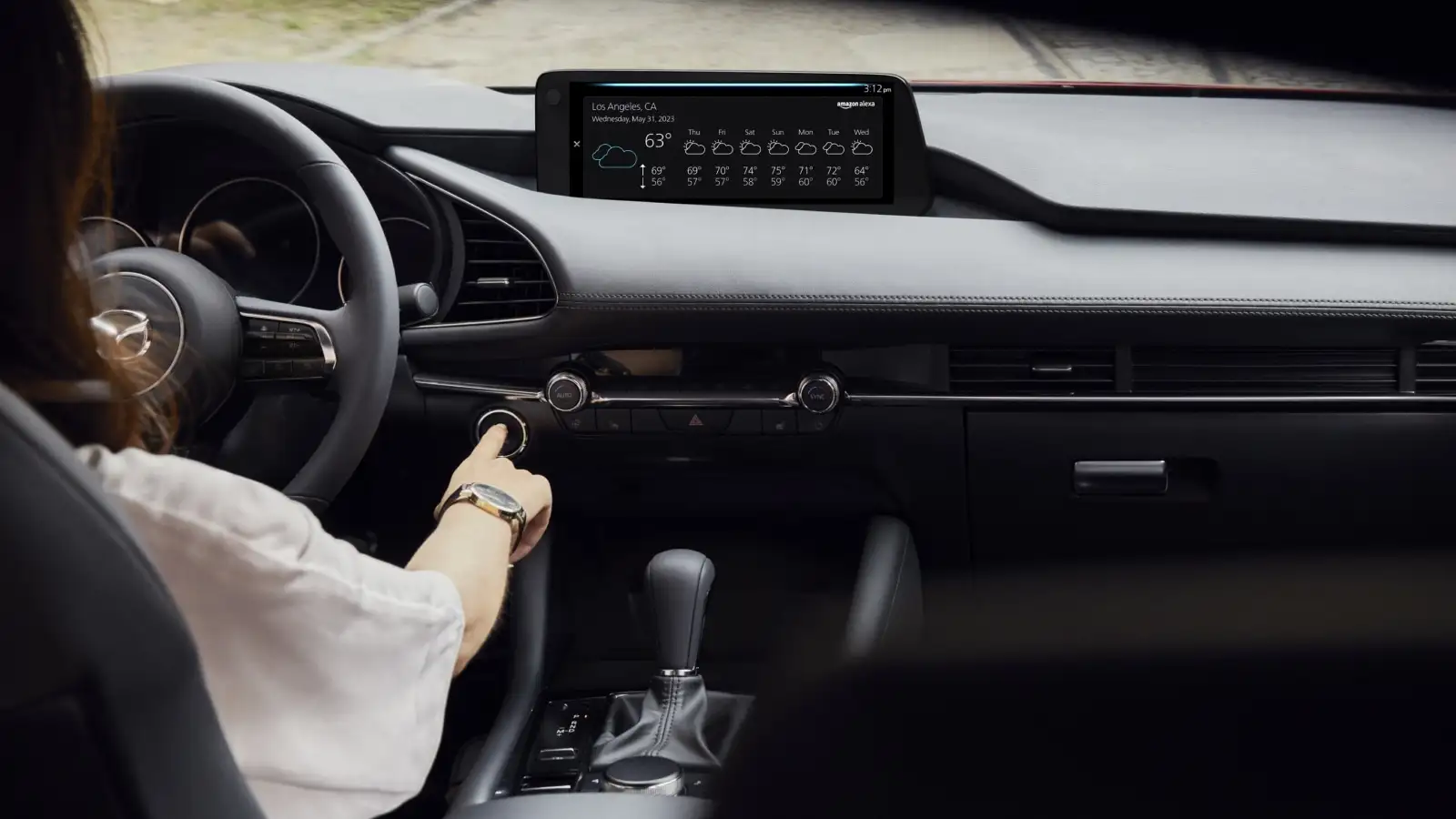 A real photograph of a car interior at dusk with a driver speaking toward the steering wheel while the center console shows an active voice assistant prompt, news photography style